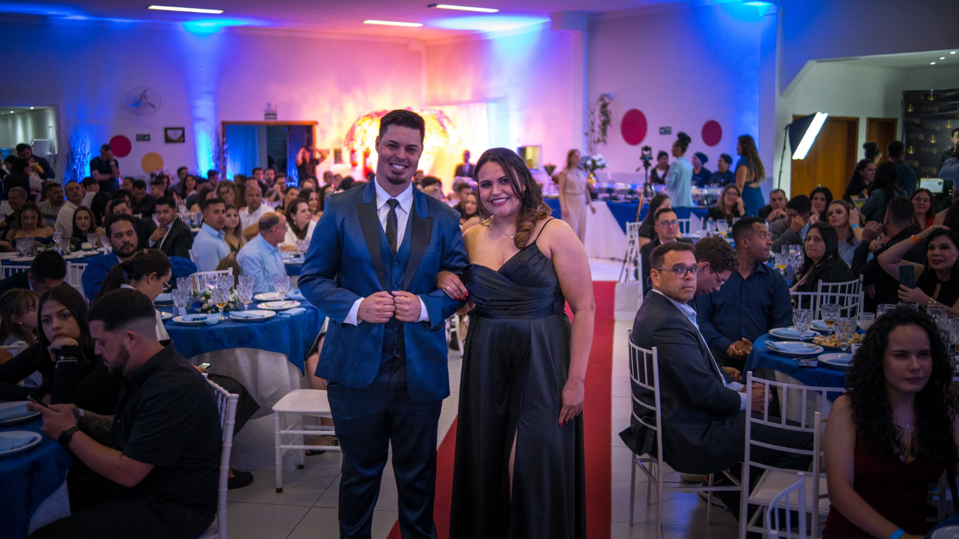 Casal em evento formal coberto pelo Ibiúna Cast, com iluminação azul e rosa ao fundo